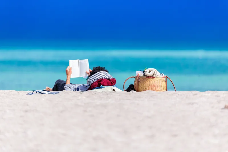 Someone lying on white sand reading a paperback with bright turquoise sea behind.