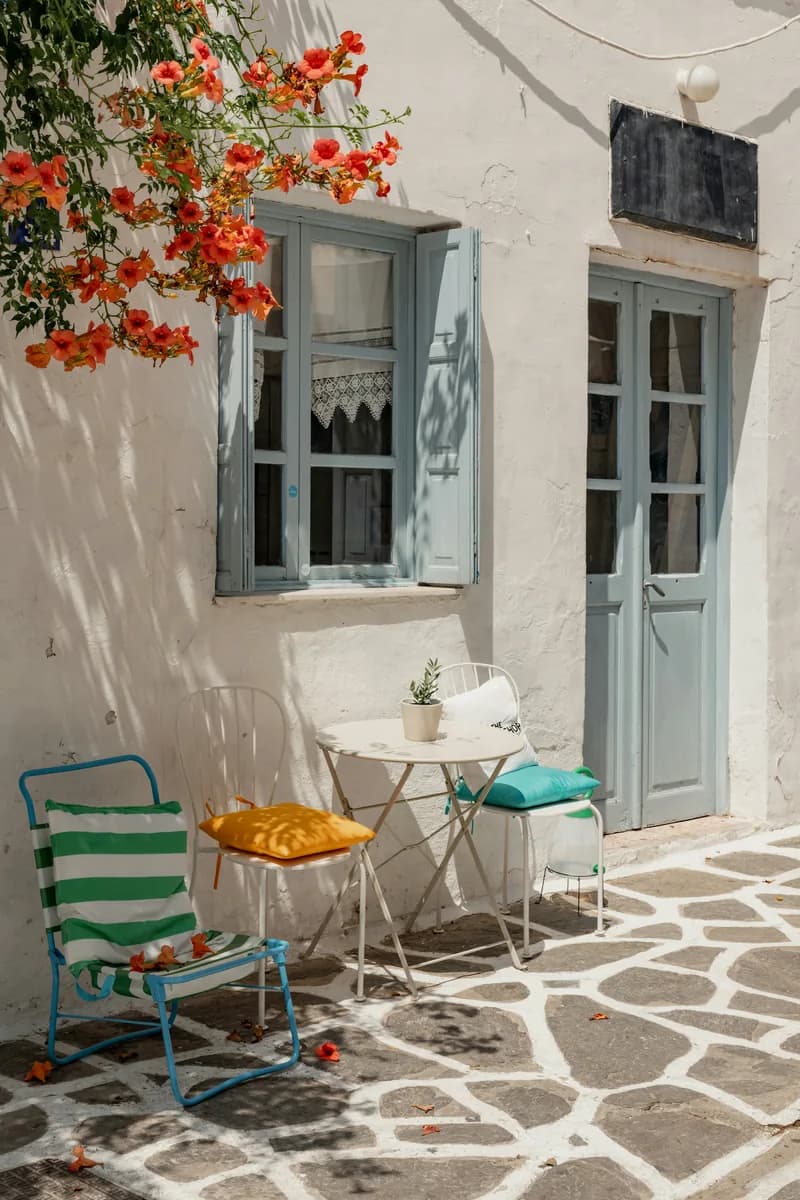 Small bistro table and mismatched chairs under flowering bougainvillea on a whitewashed lane.
